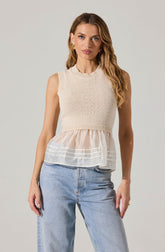 A woman is wearing a sleeveless, cream-colored knit top with a round neckline featuring a subtle ribbed pattern on the hem and armholes. The top has a layered design with a sheer, lightweight fabric extending below the knit section, creating a flowy, ruffled appearance with horizontal white stripes. She is also wearing light-wash, high-waisted jeans with a relaxed fit.